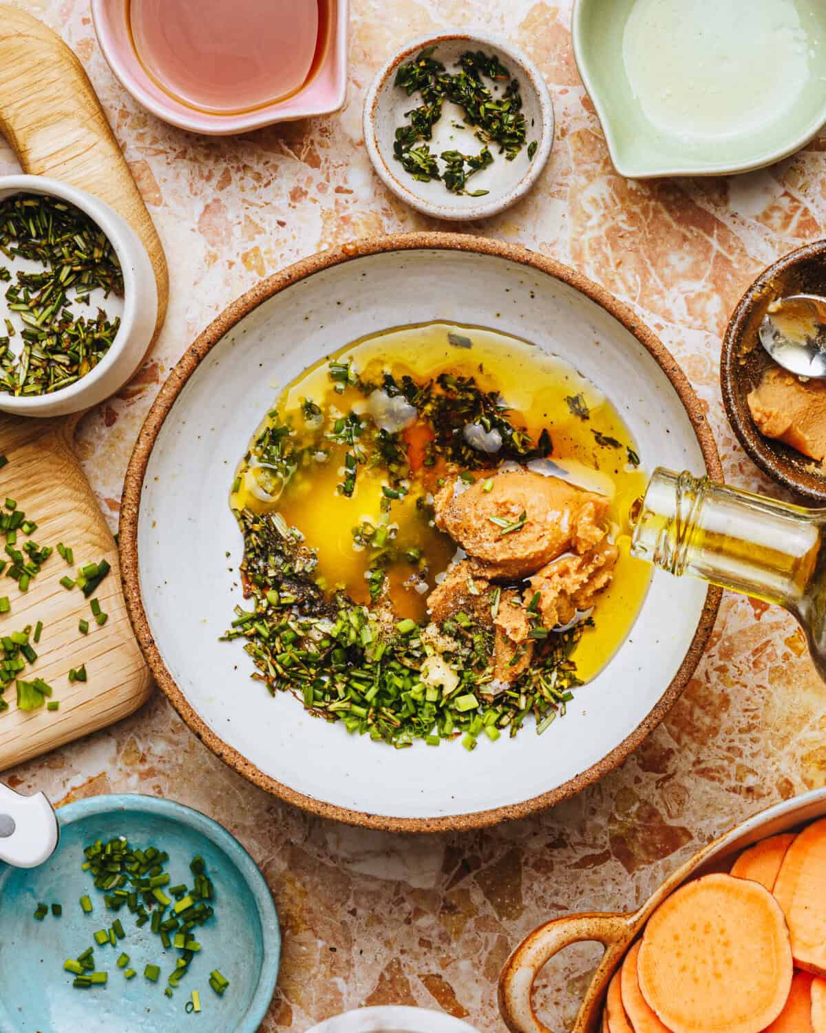 Overhead view of a bowl filled with olive oil, chopped herbs, mustard, and spices being mixed. Surrounding the bowl are small dishes and a cutting board with more herbs and ingredients on a marble counter.
