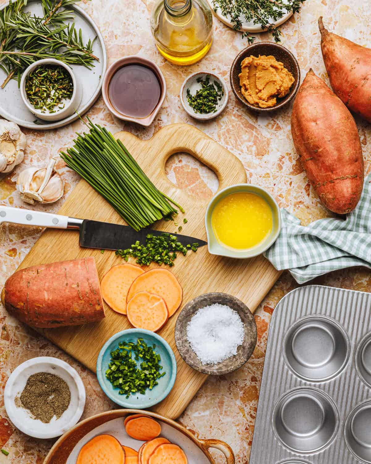 A kitchen scene with sliced sweet potatoes on a cutting board, a knife, chopped herbs, bowls of oil, salt, pepper, fresh herbs, muffin tin, and a green checkered cloth on a marble countertop.