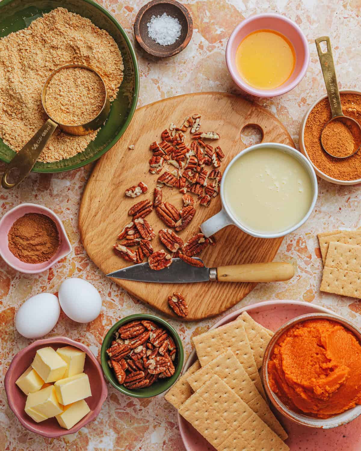 A variety of baking ingredients on a marble surface, including graham crackers, pecans, eggs, butter, brown sugar, pumpkin puree, spices, salt, melted butter, sweetened condensed milk, and a knife on a wooden board.