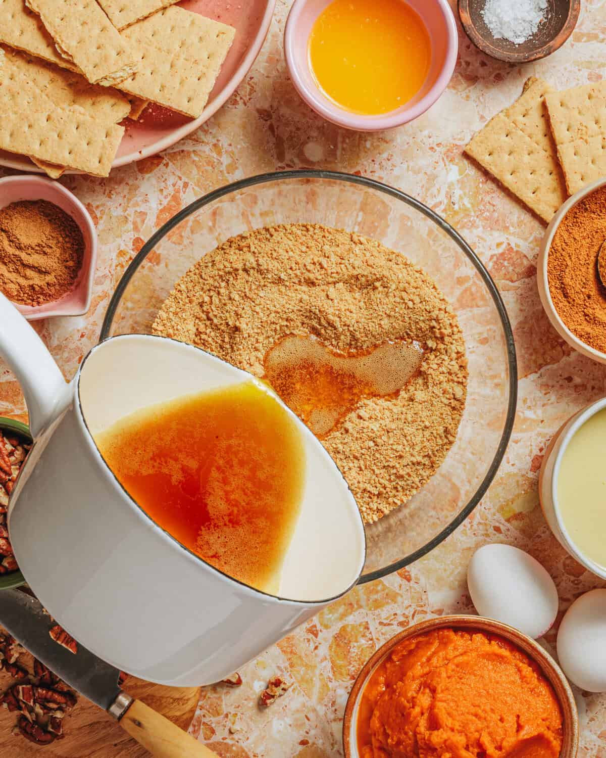A white saucepan pours melted butter into a bowl of graham cracker crumbs. Surrounding the bowl are eggs, canned pumpkin, cinnamon, condensed milk, and graham crackers on a marble countertop.
