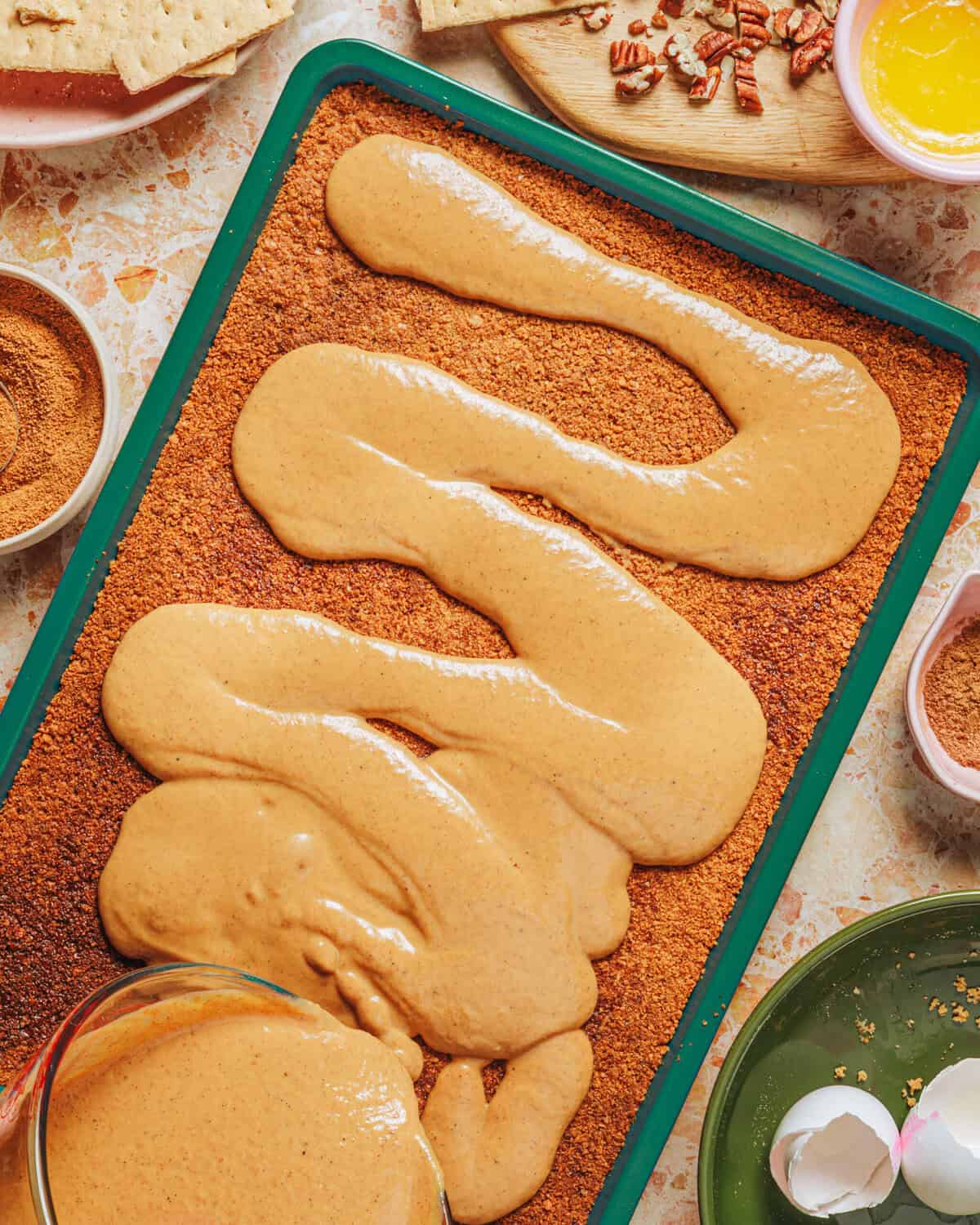 A baking pan with a graham cracker crust being covered by a thick, creamy pumpkin filling. Surrounding the pan are bowls with ingredients like cinnamon, pecans, melted butter, and broken eggshells.