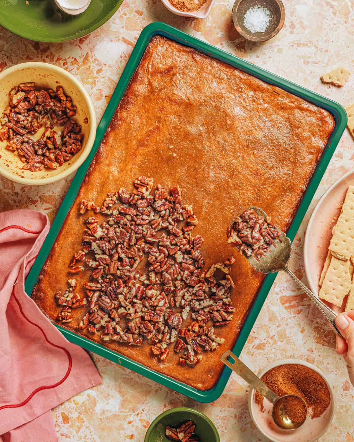 A hand sprinkles chopped pecans onto a rectangular baked dessert in a green pan, surrounded by bowls of pecans, spices, salt, and a pink cloth on a light marbled surface.