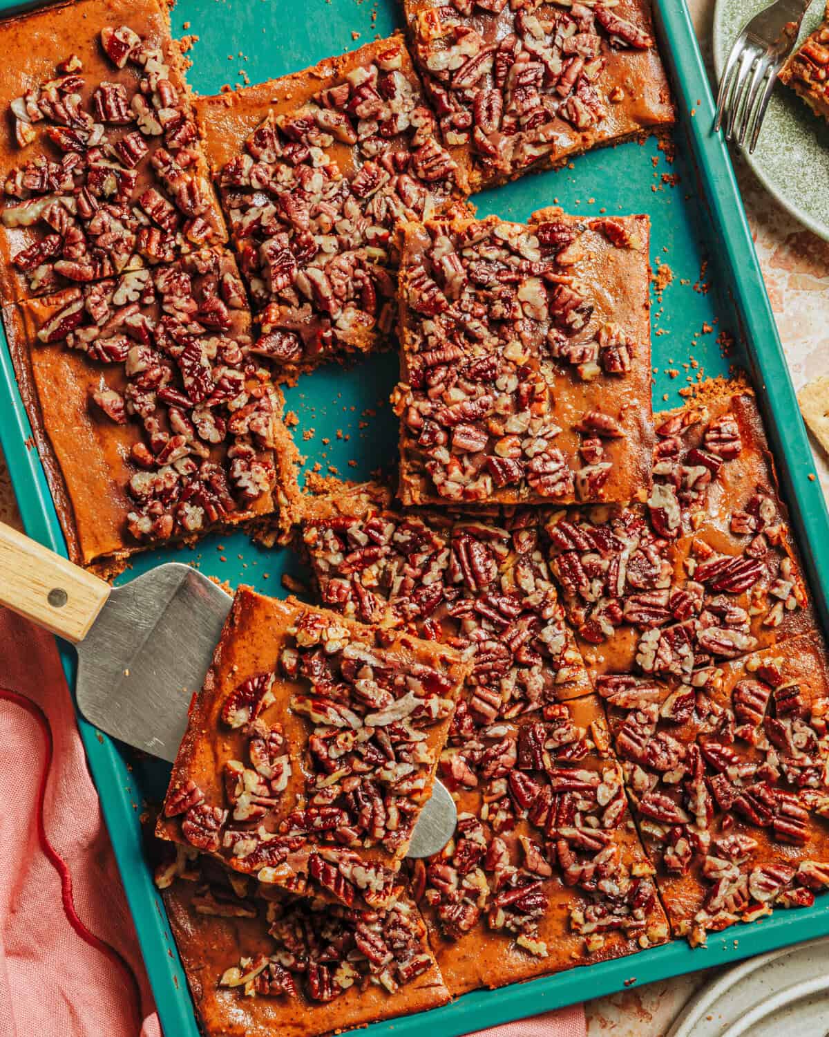 A teal baking tray holds several rectangular pecan bars with golden crust and pecan topping. A metal spatula is lifting one bar; a pink cloth and a plate are nearby on the table.