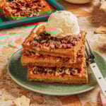 Three thick pecan bars stacked on a green plate, topped with a scoop of vanilla ice cream. A fork is beside the bars, and more pecan bars and dessert items are in the background on a speckled countertop.