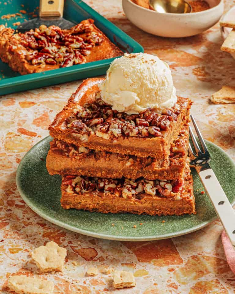 Three thick pecan bars stacked on a green plate, topped with a scoop of vanilla ice cream. A fork is beside the bars, and more pecan bars and dessert items are in the background on a speckled countertop.
