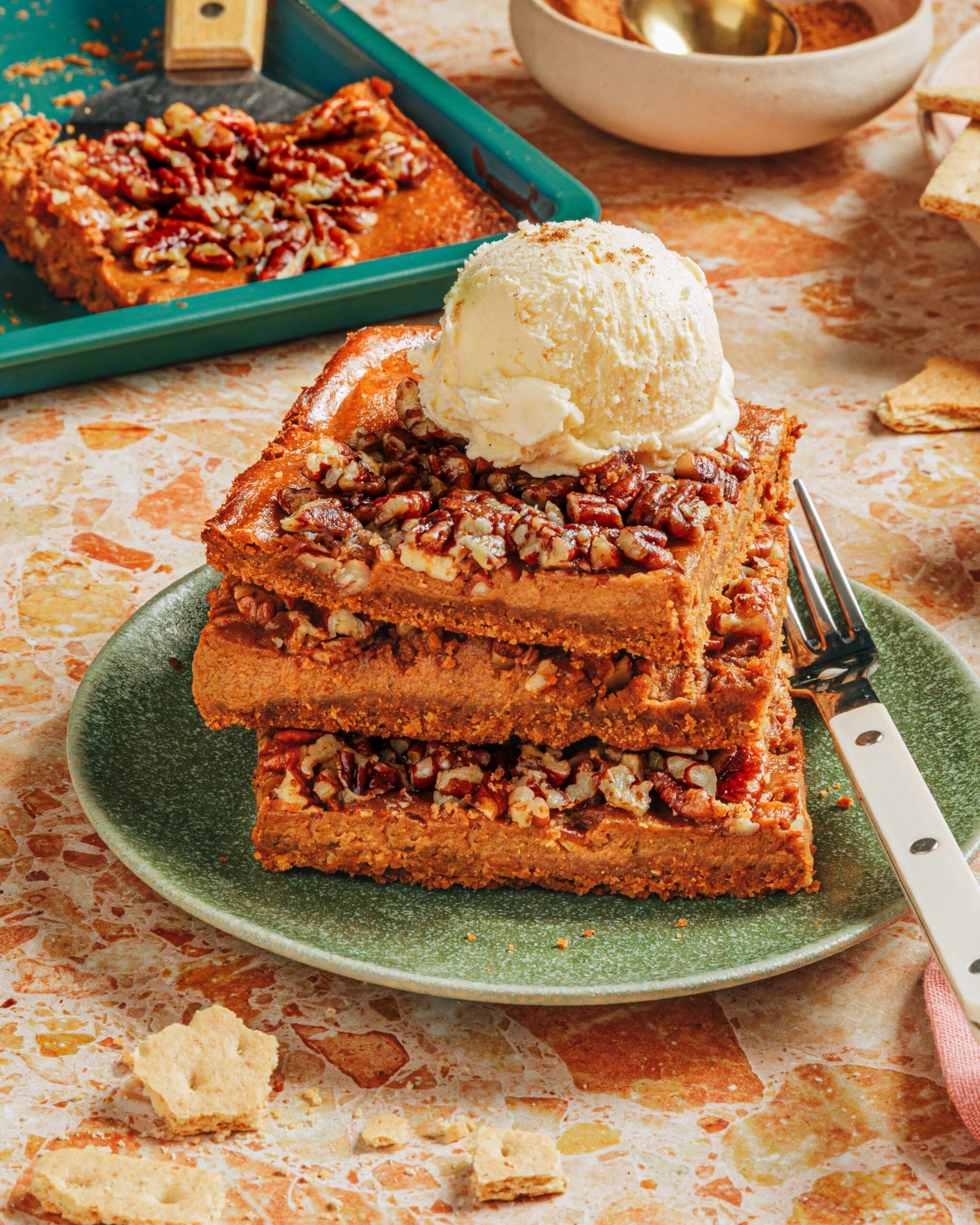 Three thick pecan bars stacked on a green plate, topped with a scoop of vanilla ice cream. A fork is beside the bars, and more pecan bars and dessert items are in the background on a speckled countertop.