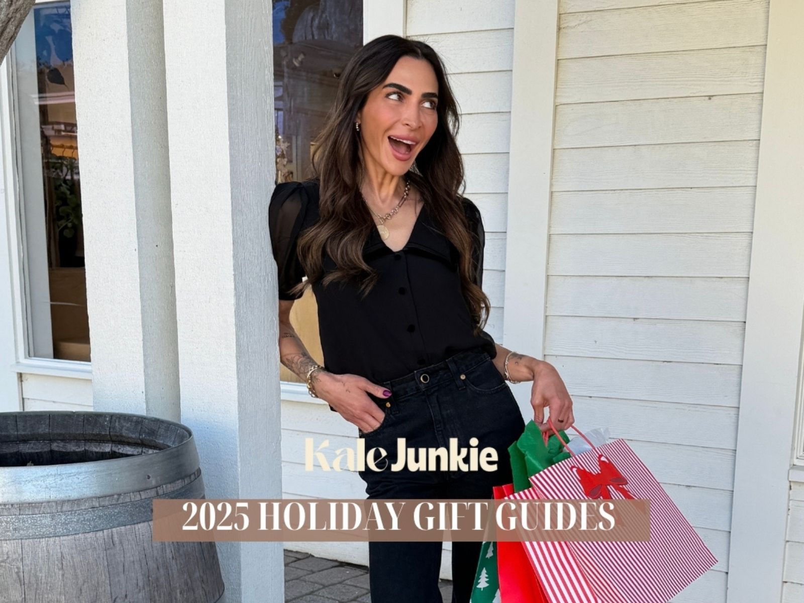 A woman with long brown hair, wearing a black blouse and dark jeans, smiles while holding holiday gift bags outside a white building. Text reads, "Kale Junkie 2025 Holiday Gift Guides.