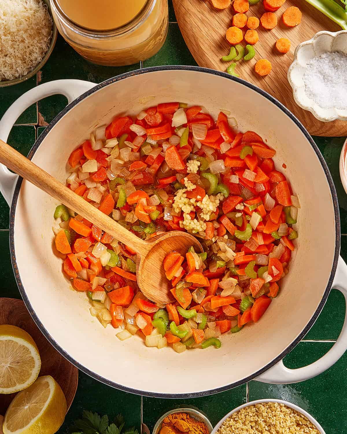 A white pot on a green countertop contains chopped carrots, celery, onions, and minced garlic being stirred with a wooden spoon. Surrounding the pot are sliced vegetables, a jar of broth, lemon halves, and salt.