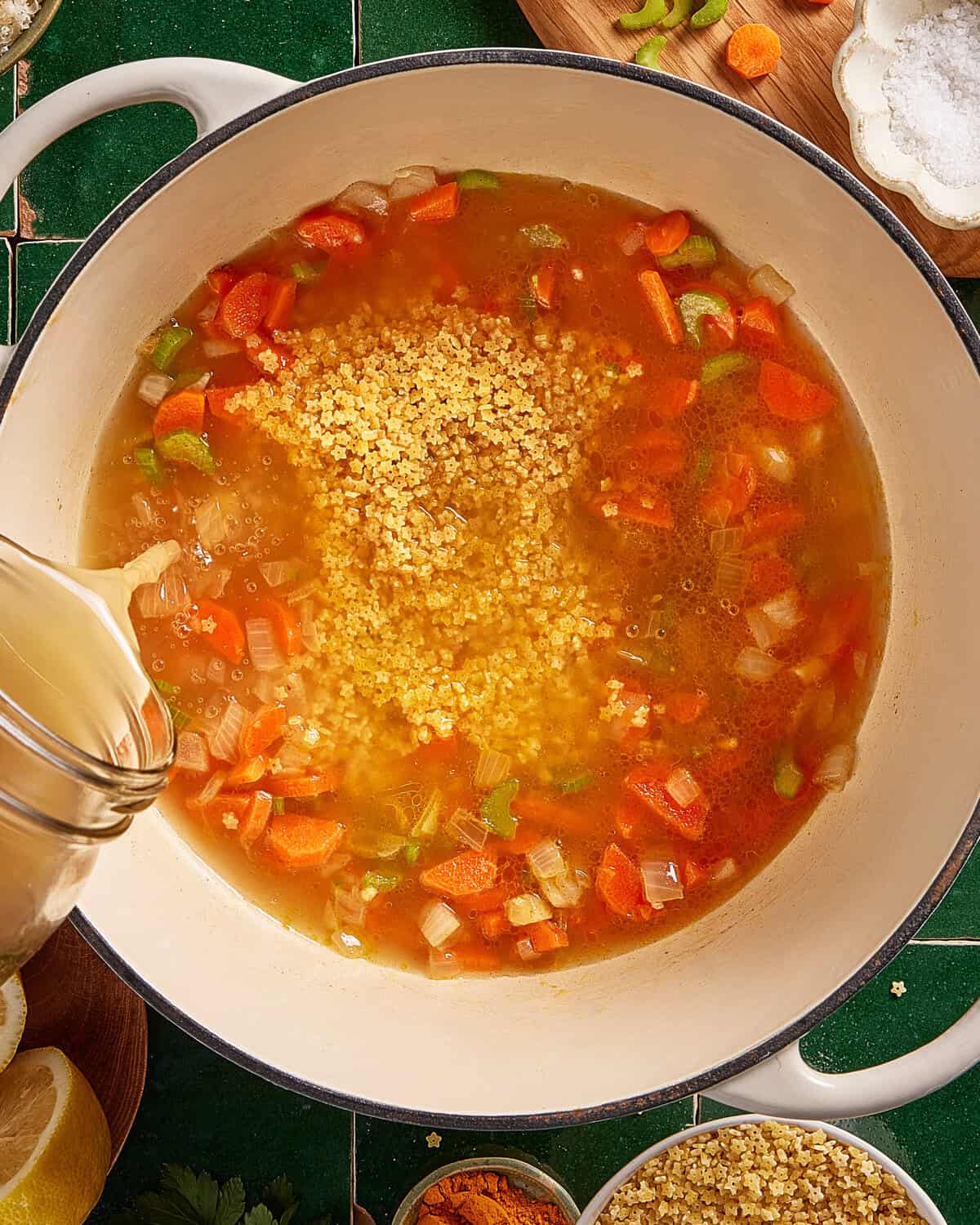 A pot of soup with diced carrots, celery, onions, and dry quinoa being poured with broth. Ingredients and seasonings are arranged around the pot on a green tiled surface.