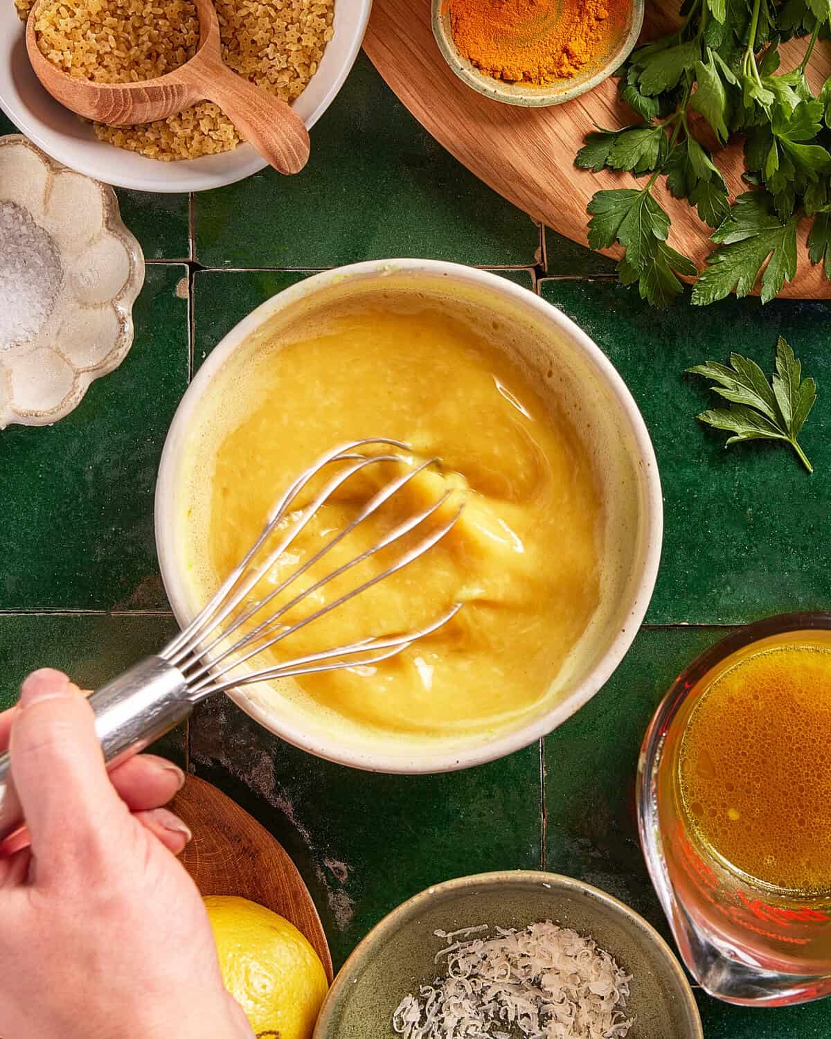 A hand whisks beaten eggs in a bowl on a green tiled surface, surrounded by parsley, grated cheese, lemon, salt, a wooden spoon with brown sugar, and a cup of yellow liquid.
