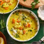A bowl of creamy soup with rice, carrots, celery, and herbs, topped with grated cheese. Fresh parsley, a lemon wedge, and a small bowl of salt sit nearby on a green tiled surface.