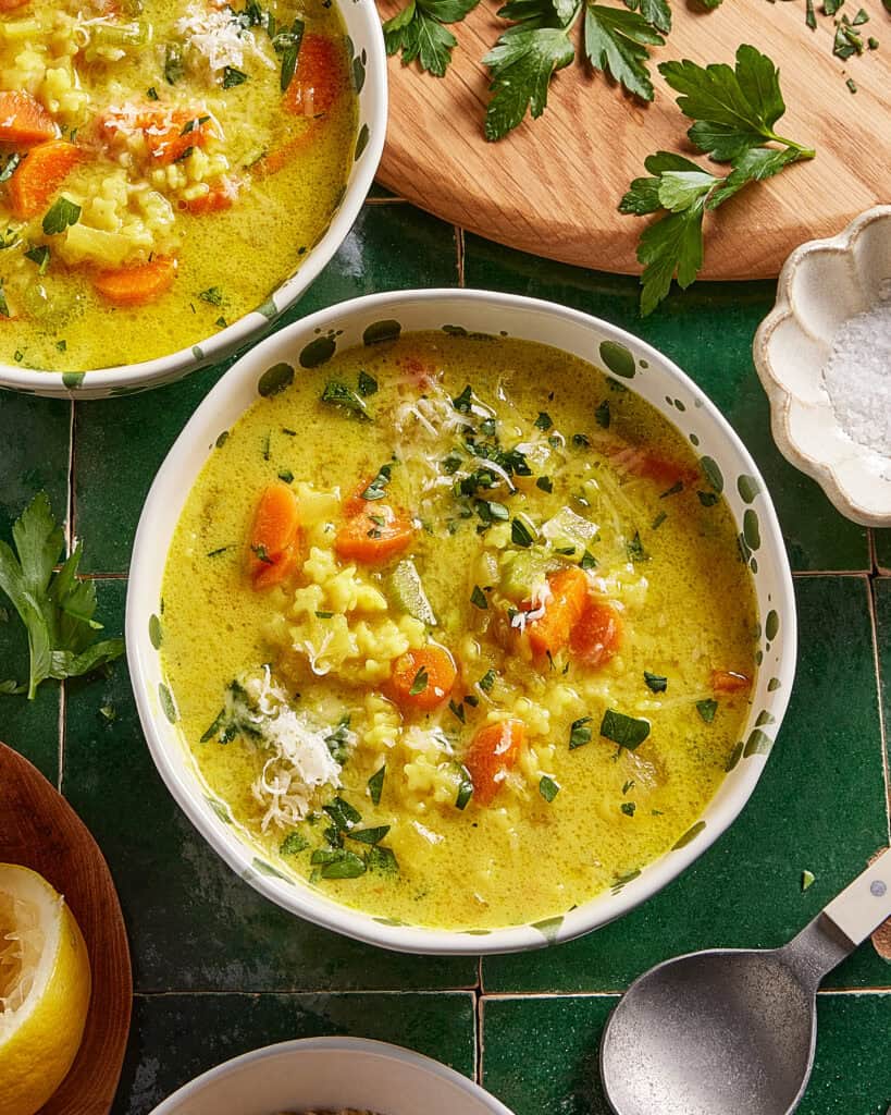 A bowl of creamy soup with rice, carrots, celery, and herbs, topped with grated cheese. Fresh parsley, a lemon wedge, and a small bowl of salt sit nearby on a green tiled surface.