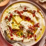 A bowl of creamy mashed cauliflower topped with melted butter, crispy bacon bits, and chopped herbs, with a spoon resting on the side. Raw cauliflower florets and a pink napkin are nearby.