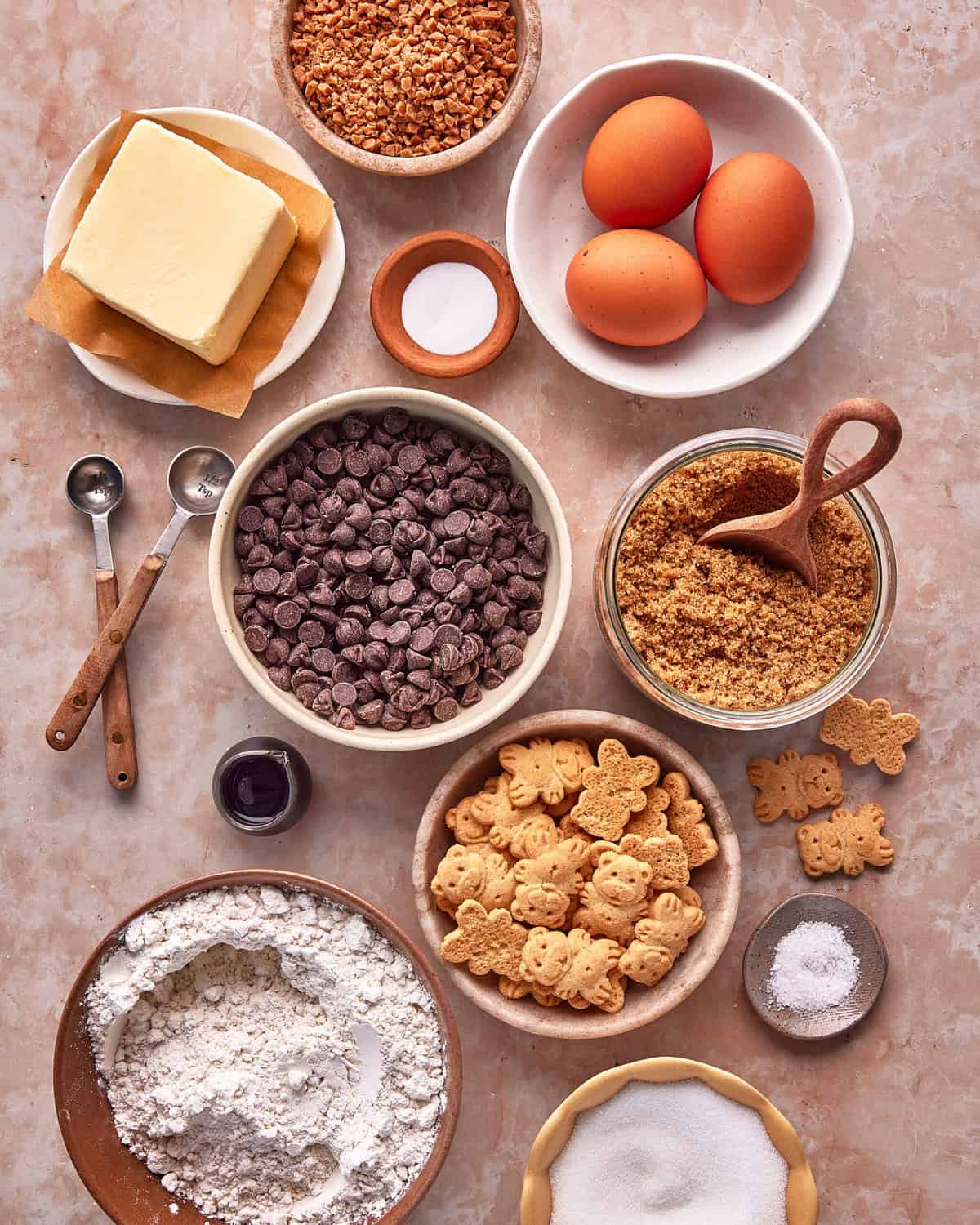 Baking ingredients on a countertop, including butter, eggs, chocolate chips, brown sugar, flour, granulated sugar, mini bear cookies, chopped nuts, salt, measuring spoons, and a small bowl of vanilla extract.