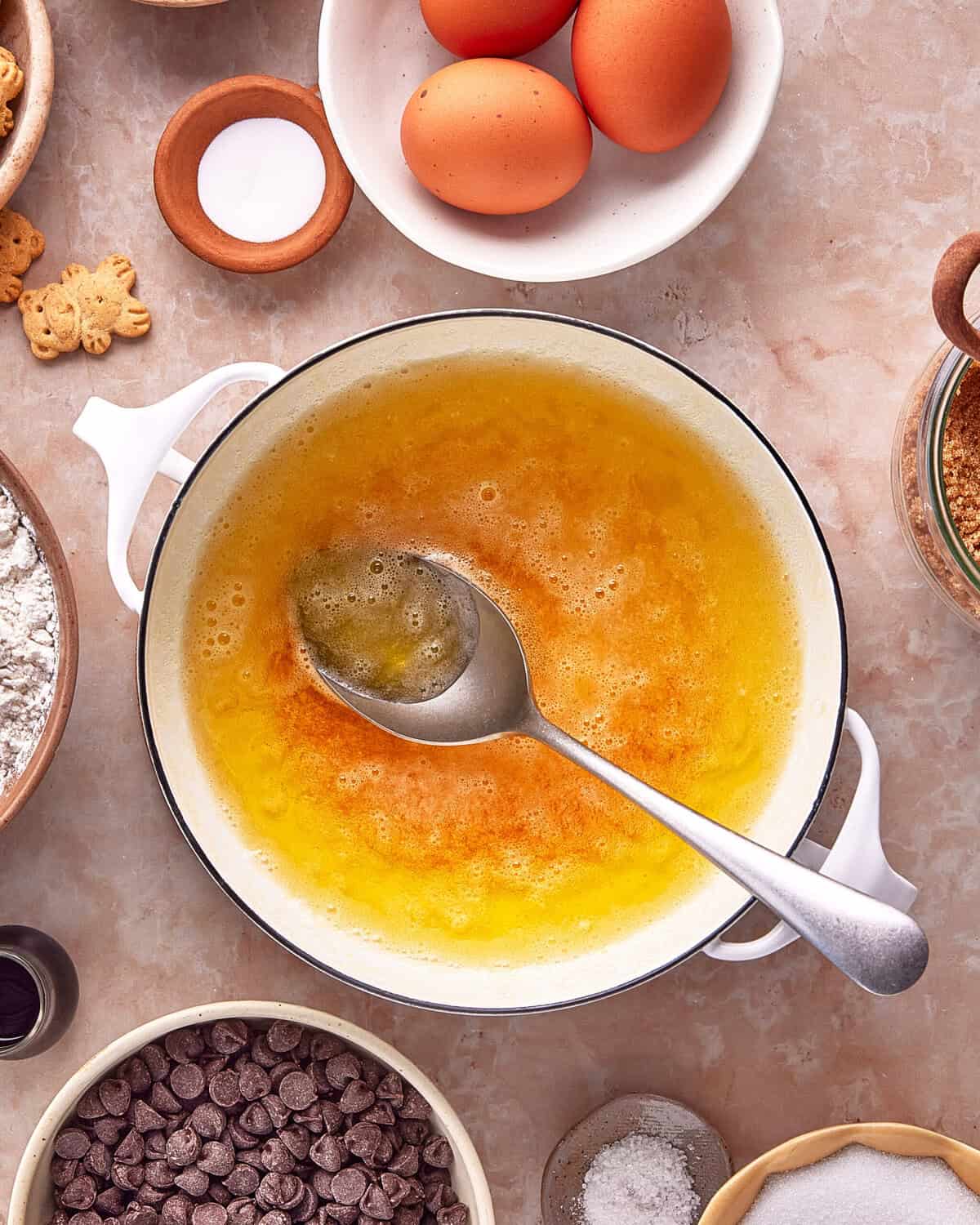 Melted butter is being stirred with a spoon in a white pot, surrounded by eggs, flour, chocolate chips, animal crackers, sugar, salt, and vanilla on a light countertop.