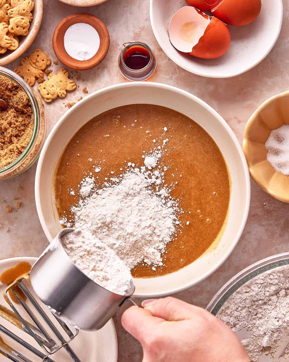 A hand pours flour from a measuring cup into a mixing bowl of batter. Surrounding the bowl are baking ingredients, including eggs, sugar, vanilla, brown sugar, cookies, and salt on a countertop.