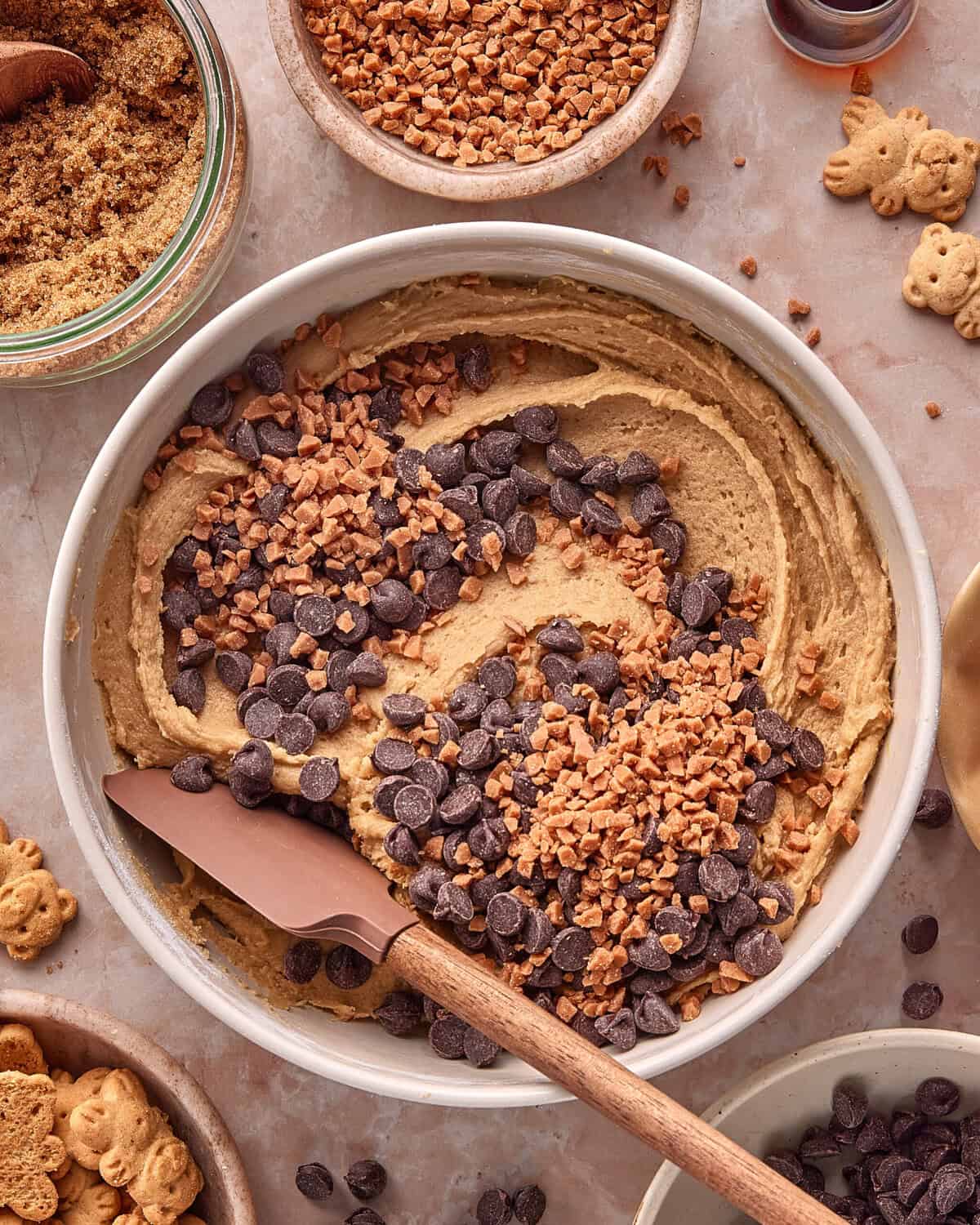 A mixing bowl filled with cookie dough, chocolate chips, and toffee bits, with a spatula resting inside. Surrounding the bowl are small dishes of brown sugar, toffee bits, vanilla, and bear-shaped cookies.