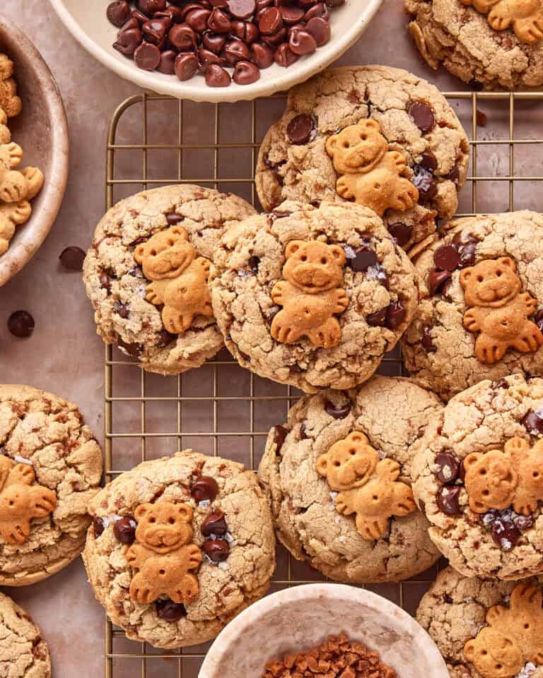 Chocolate chip cookies topped with small bear-shaped biscuits are arranged on a cooling rack. Bowls of chocolate chips and other ingredients surround the cookies.