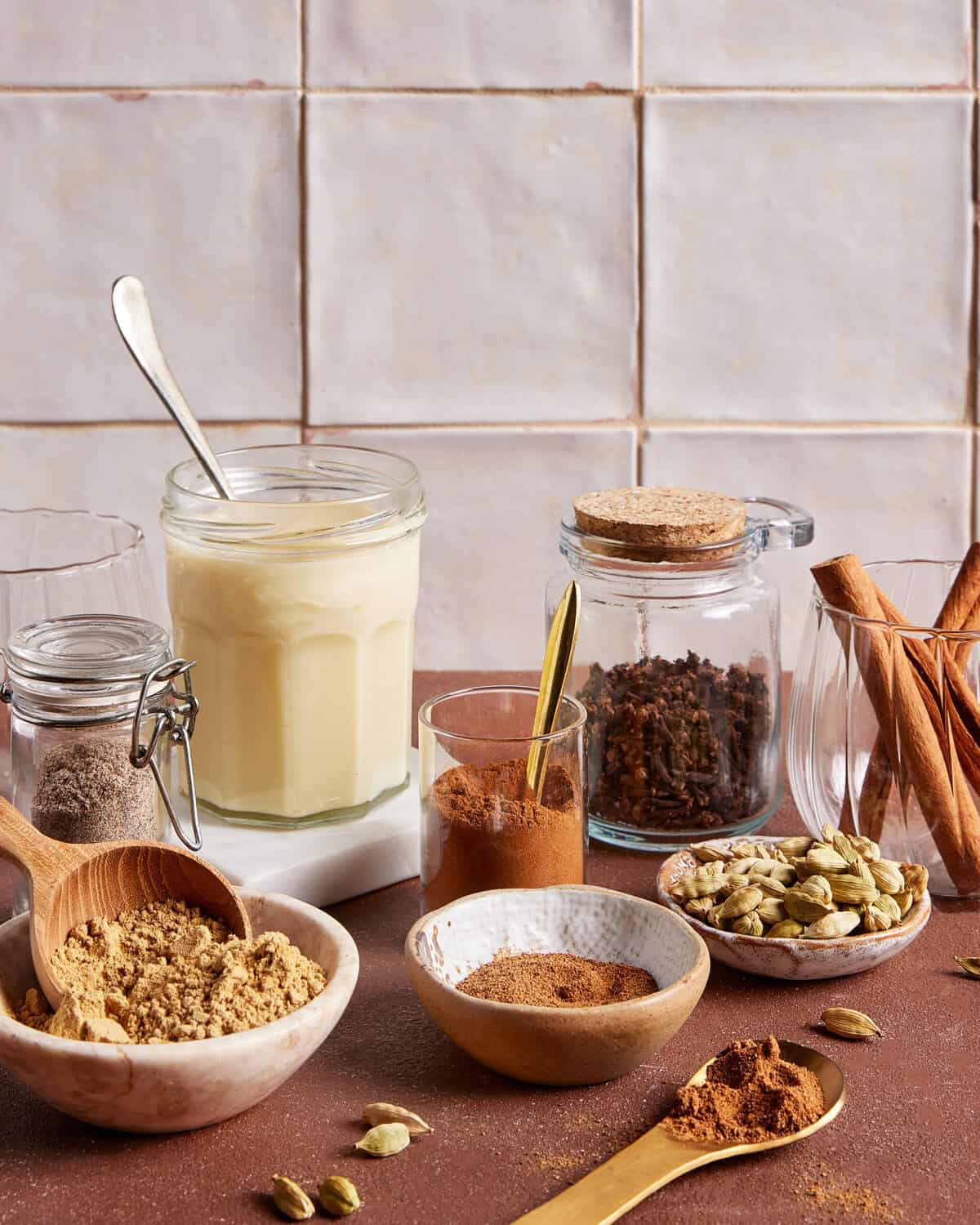 A variety of spices and ingredients on a kitchen counter, including jars of ground spices, a glass of creamy liquid, bowls of powders, whole cardamom pods, cinnamon sticks, and a wooden spoon.