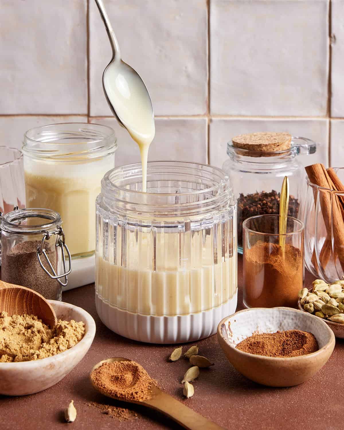 A spoon drizzles condensed milk into a glass jar, surrounded by jars and bowls filled with spices like cinnamon, ground ginger, cardamom pods, and brown sugar on a brown countertop with tiled background.