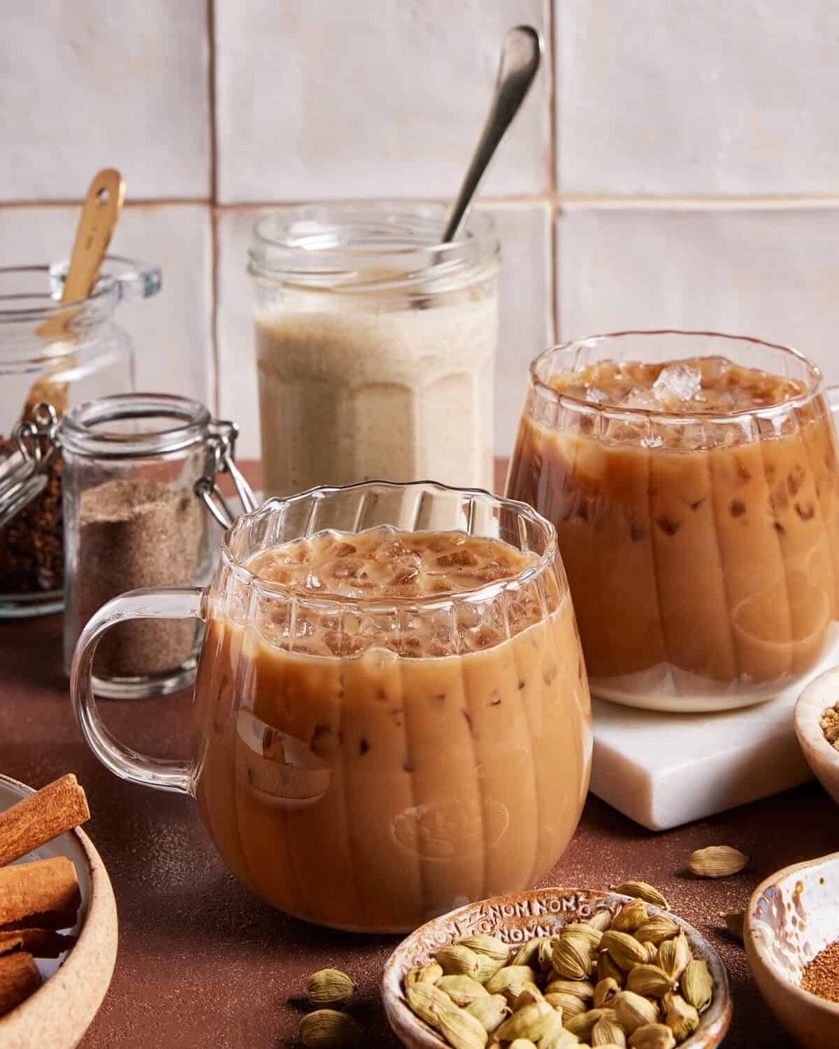 Two clear glass mugs filled with iced chai latte sit on a table, surrounded by spices like cinnamon sticks and cardamom pods, with jars of chai concentrate and spice mix in the background.