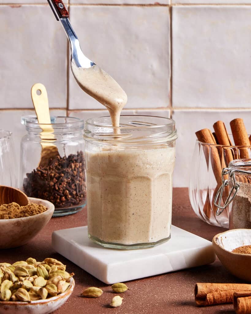 A spoon drizzles creamy chai-spiced spread into a glass jar. Around it are jars of spices, cinnamon sticks, whole cardamom pods, cloves, and a bowl of brown sugar on a brown surface with a tiled background.