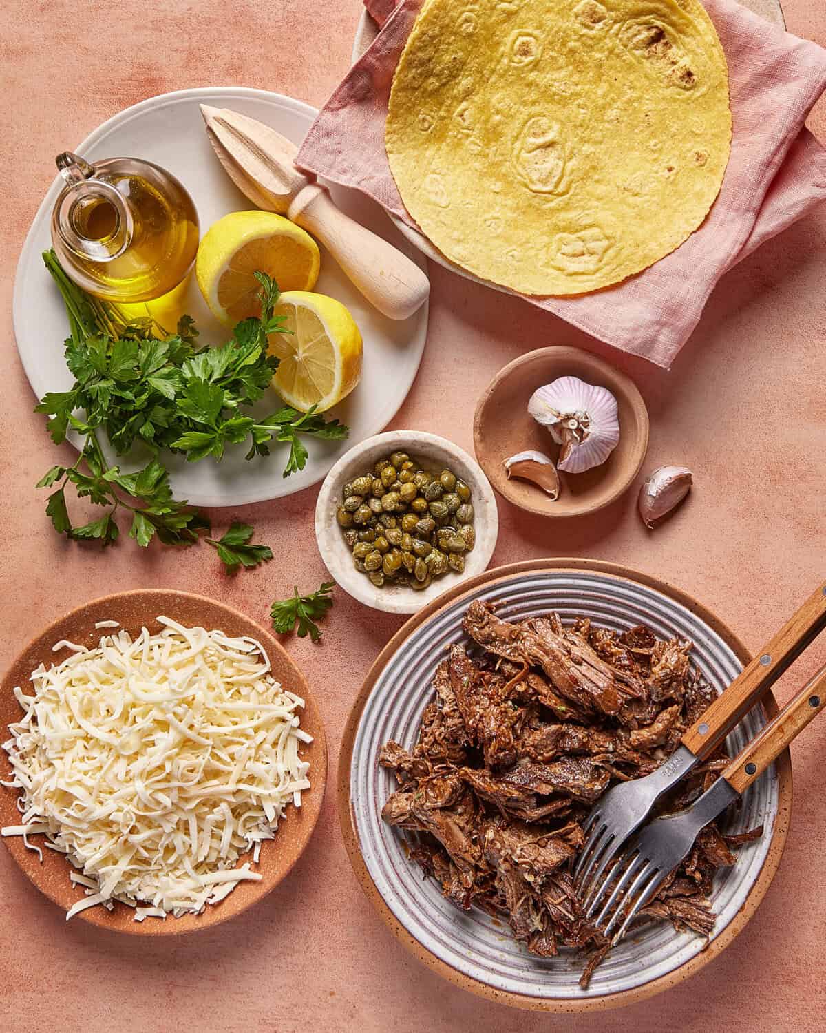A flat lay of shredded beef on a plate with forks, grated cheese, fresh parsley, lemon halves, capers, garlic, olive oil, and a corn tortilla on a cloth napkin, all on a pinkish surface.