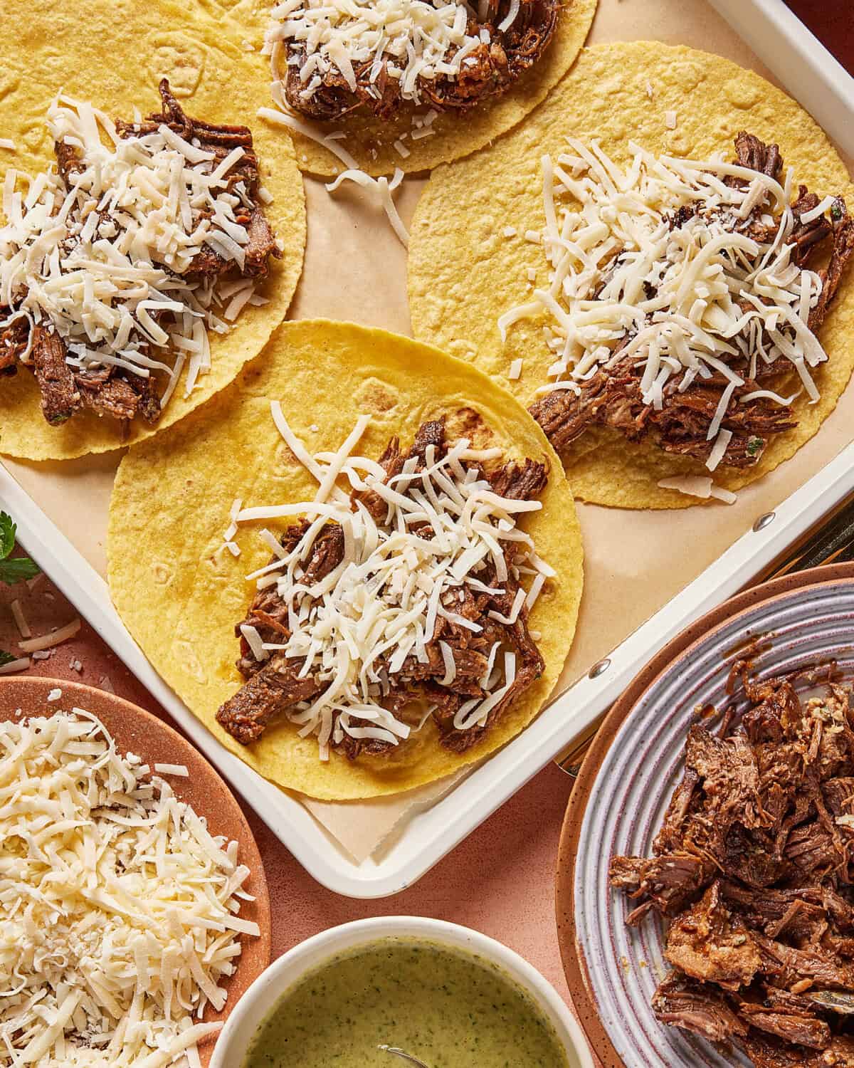 Five corn tortillas topped with shredded beef and grated cheese are arranged on a tray. Surrounding dishes hold extra shredded beef, grated cheese, and a bowl of green sauce.