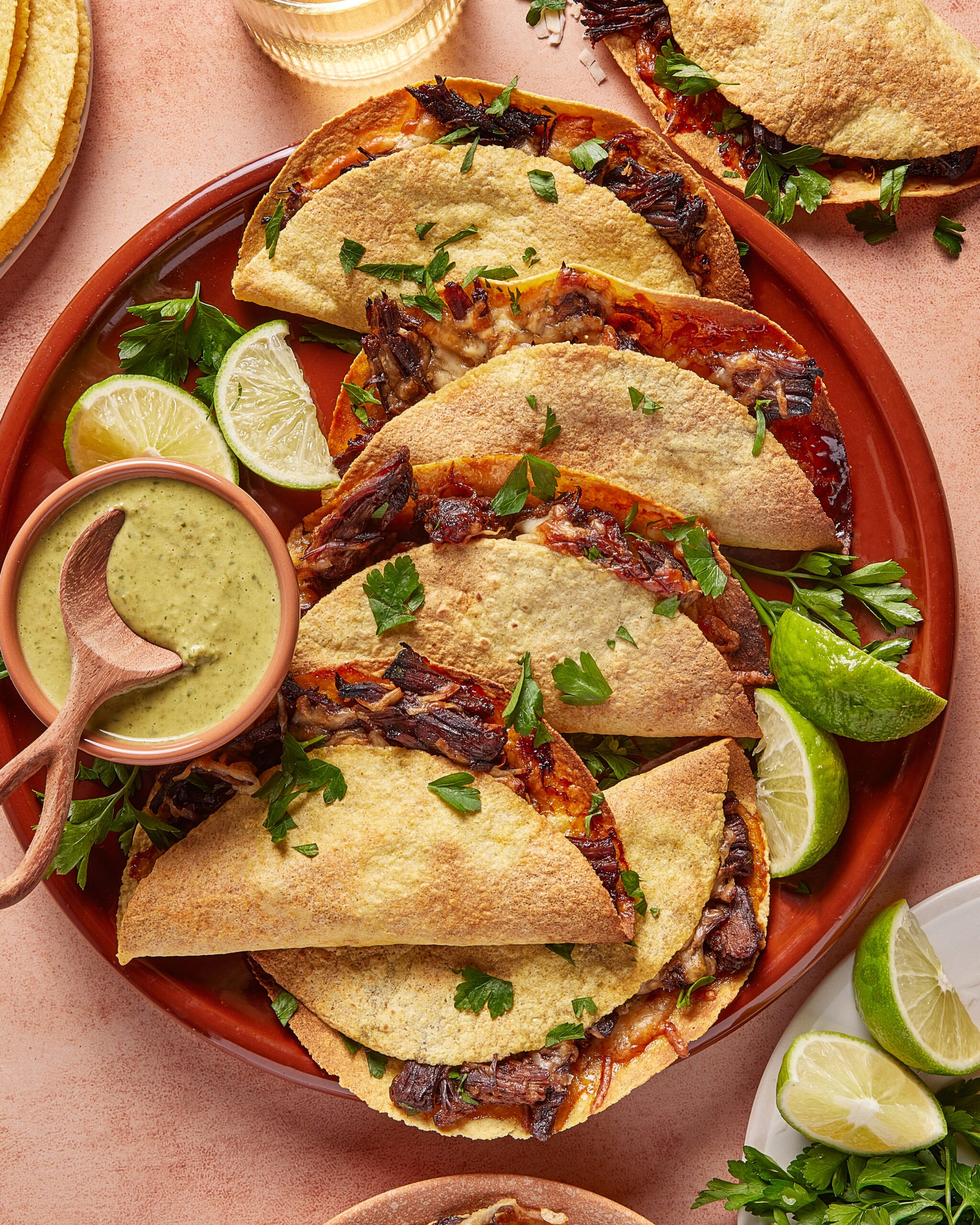A plate of folded tacos with grilled vegetables and fresh herbs, served with lime wedges and a small bowl of green sauce, set on a terra-cotta colored surface.