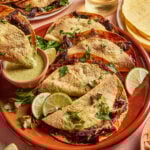 A plate of crispy tacos filled with shredded meat, garnished with parsley and lime wedges. A small dish of green sauce is on the side, and there are more tacos, corn tortillas, and a glass of drink in the background.
