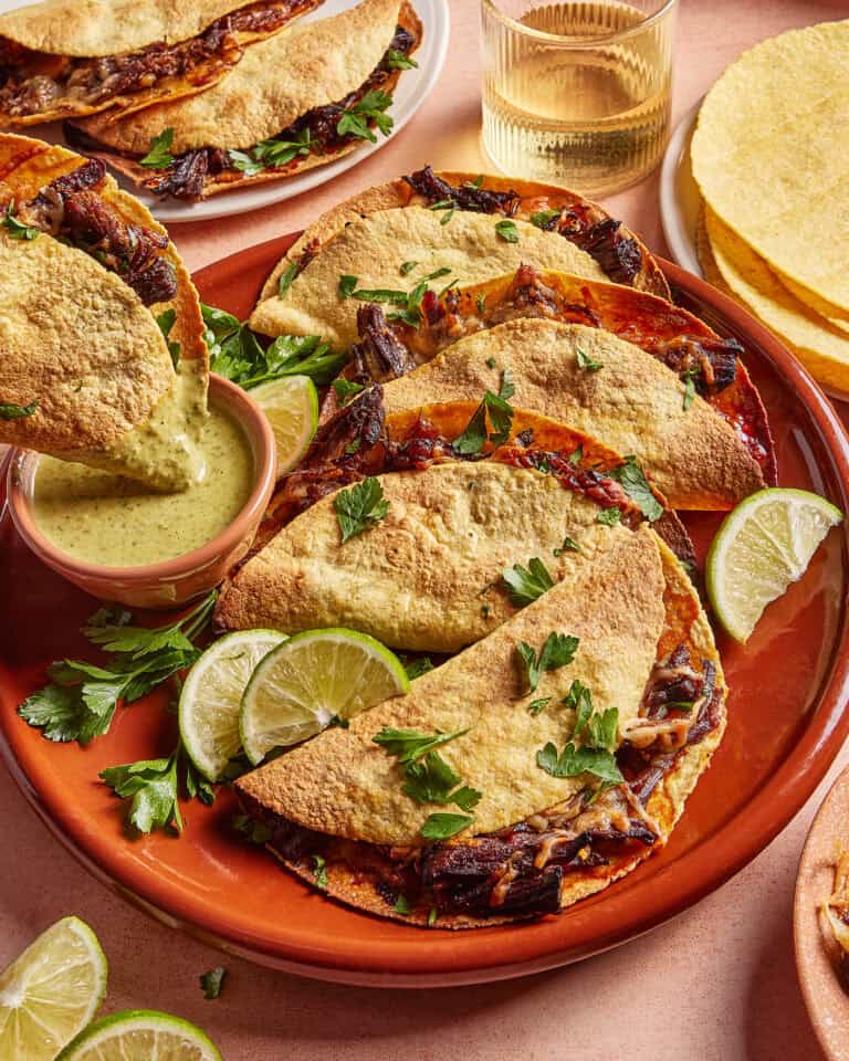 A plate of crispy tacos filled with shredded meat, garnished with parsley and lime wedges. A small dish of green sauce is on the side, and there are more tacos, corn tortillas, and a glass of drink in the background.
