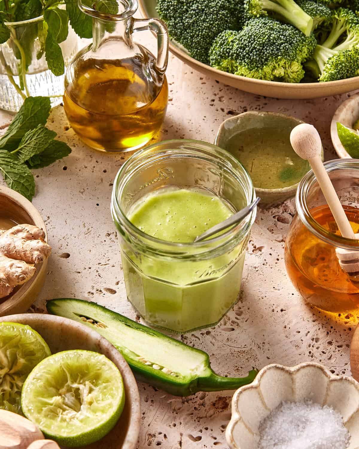 A jar of green smoothie with a spoon sits on a textured surface, surrounded by fresh ingredients like cucumber, lime, broccoli, ginger, mint, honey, olive oil, and a bowl of salt.