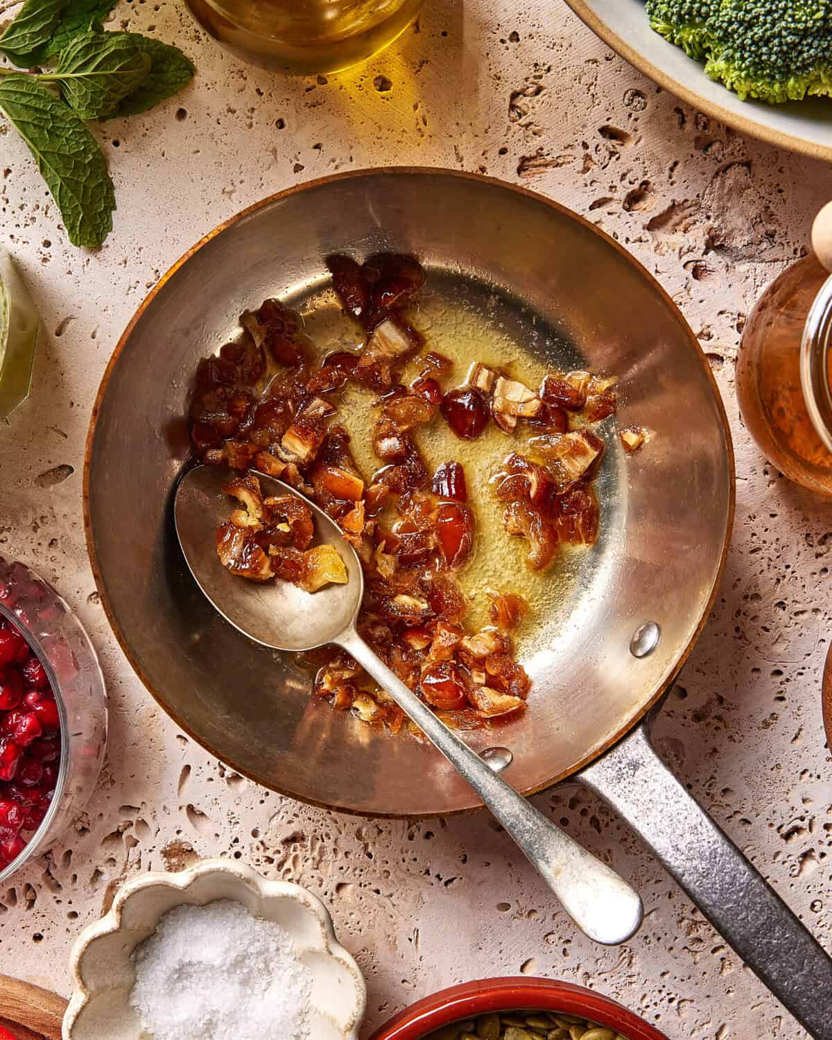 A metal pan with chopped dates being sautéed in oil, stirred by a silver spoon. Surrounding the pan are bowls of pomegranate seeds, salt, broccoli, herbs, and honey on a textured surface.