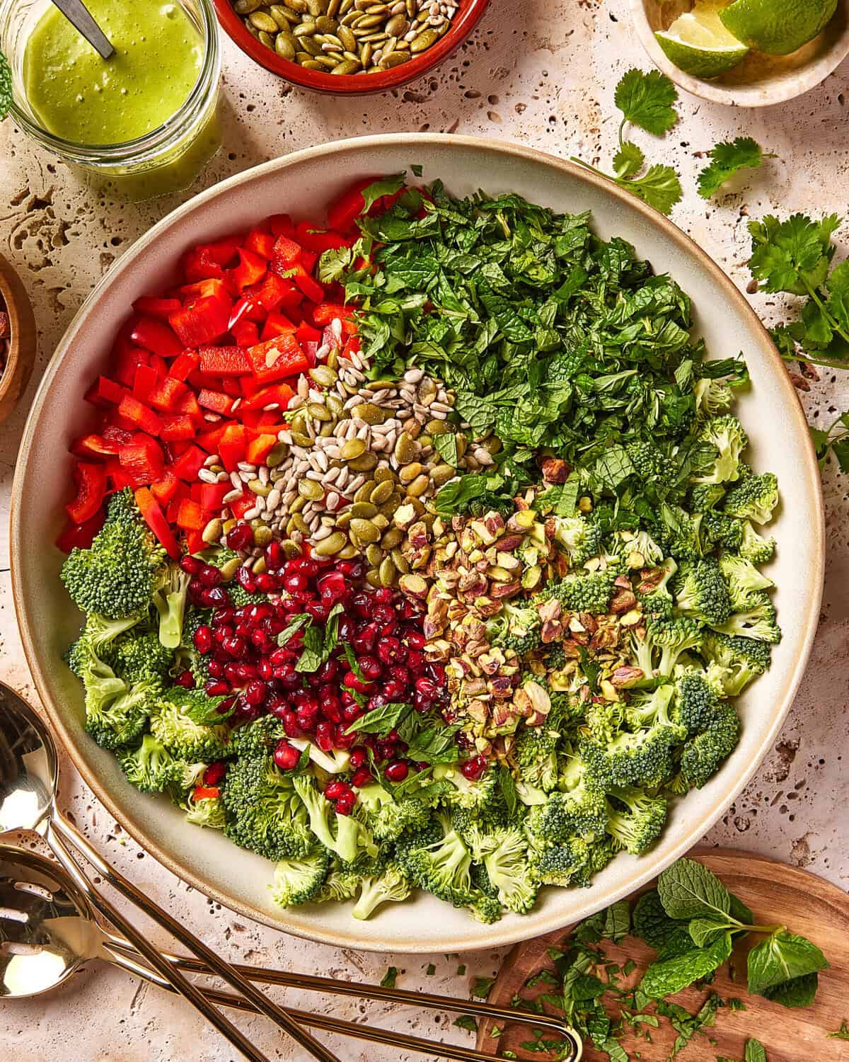 A large bowl contains chopped red bell pepper, broccoli florets, mint, parsley, pomegranate seeds, sunflower seeds, pepitas, and pistachios, all arranged in separate, colorful sections. Surrounding the bowl are various ingredients.