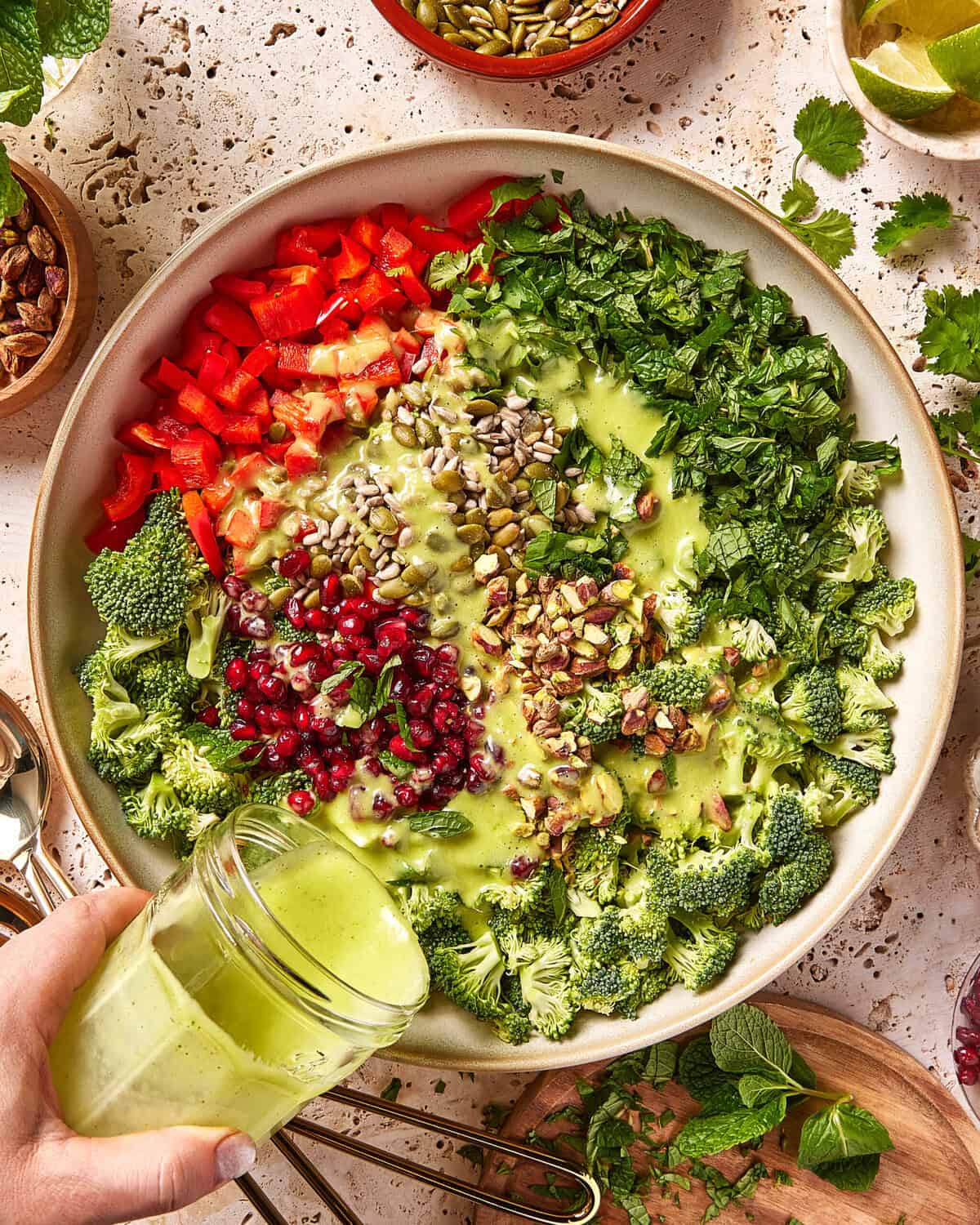 A large bowl filled with chopped broccoli, red bell pepper, mint, parsley, pomegranate seeds, sunflower seeds, and pumpkin seeds, with green dressing being poured over the salad.