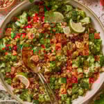 A vibrant broccoli salad with chopped red bell peppers, pomegranate seeds, golden raisins, pumpkin seeds, cilantro, and lime slices, served in a bowl with two serving spoons on a textured surface.