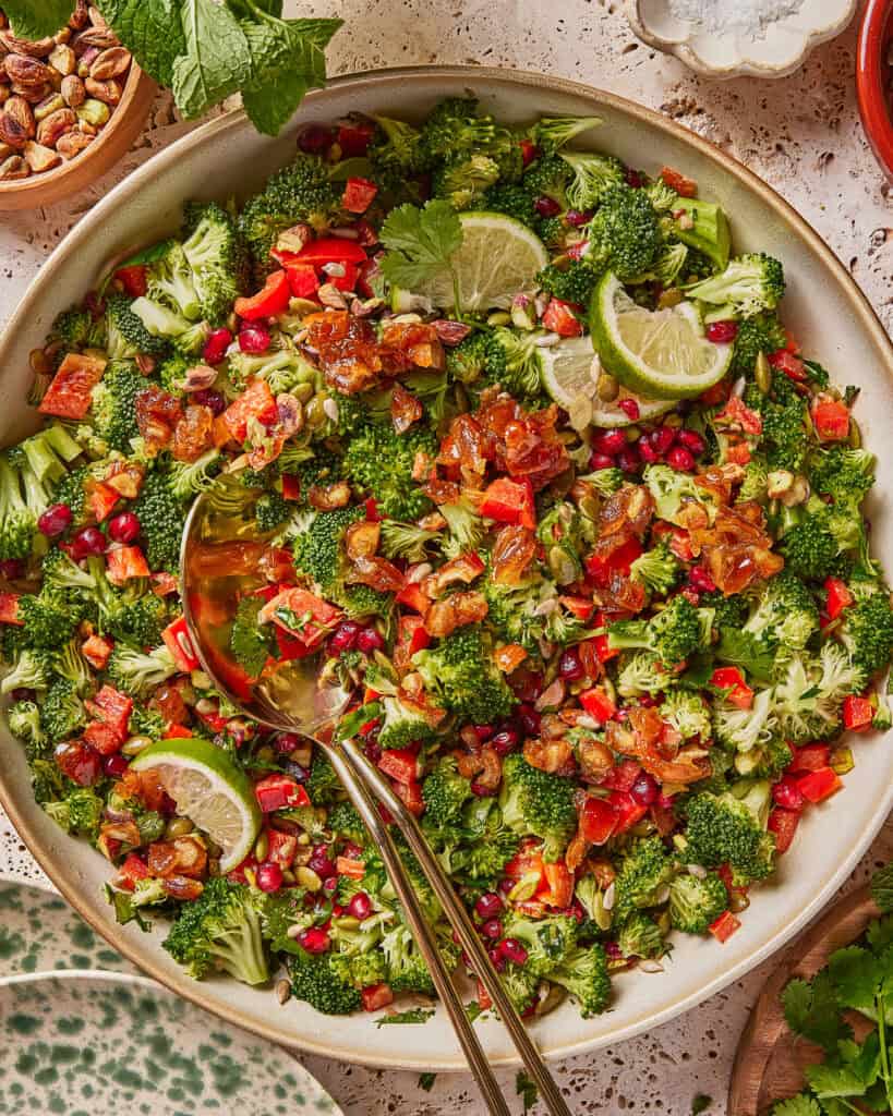 A vibrant broccoli salad with chopped red bell peppers, pomegranate seeds, golden raisins, pumpkin seeds, cilantro, and lime slices, served in a bowl with two serving spoons on a textured surface.