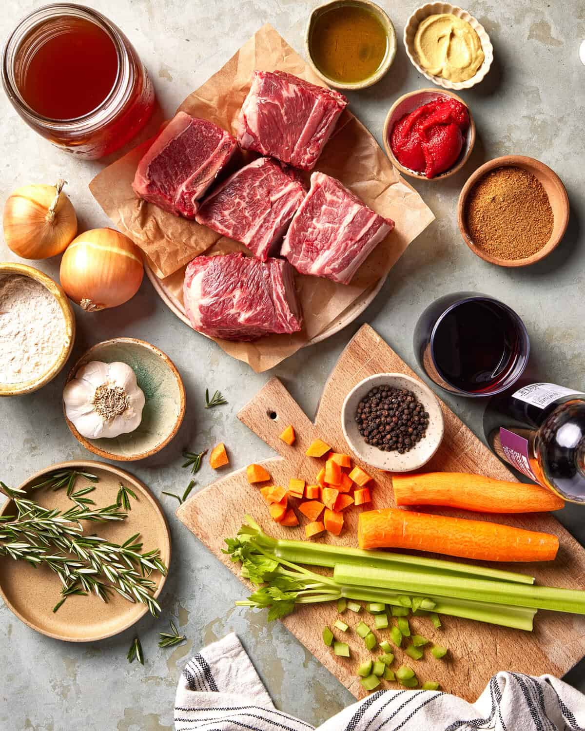 Overhead view of raw short ribs on parchment, surrounded by onions, garlic, rosemary, chopped carrots, celery, flour, black pepper, olive oil, tomato paste, mustard, broth, brown sugar, and red wine on a countertop.