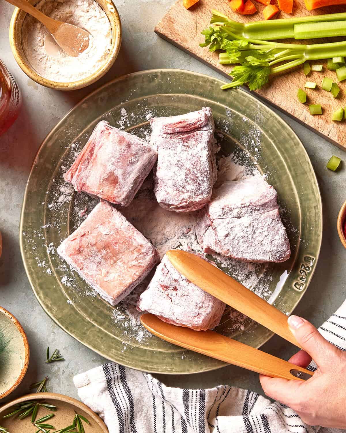 Five pieces of raw, floured meat sit on a green plate. A hand holds wooden tongs near the meat. Surrounding the plate are chopped vegetables, flour, and cooking utensils on a kitchen countertop.