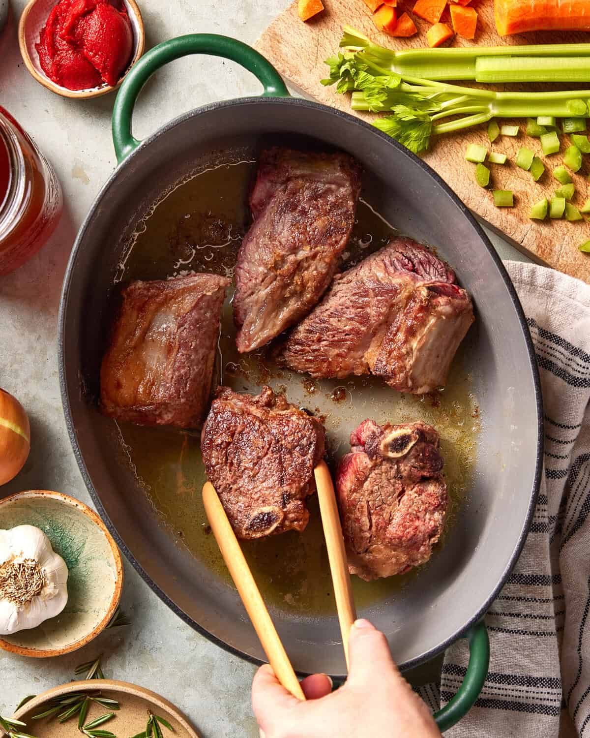 A person uses tongs to sear large beef short ribs in a gray oval pan. Surrounding the pan are chopped carrots, celery, tomato paste, garlic, onion, and rosemary on a kitchen counter.