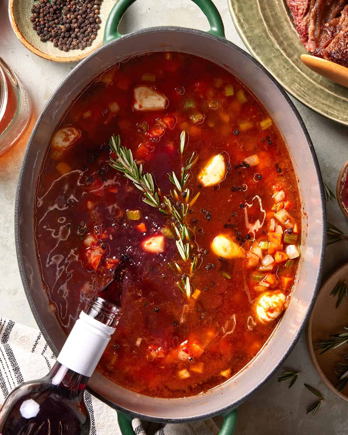 A pot of rich red stew with vegetables, garlic cloves, black peppercorns, and a rosemary sprig, while red wine is being poured in. Surrounding the pot are ingredients like black pepper, steak, and kitchen towels.