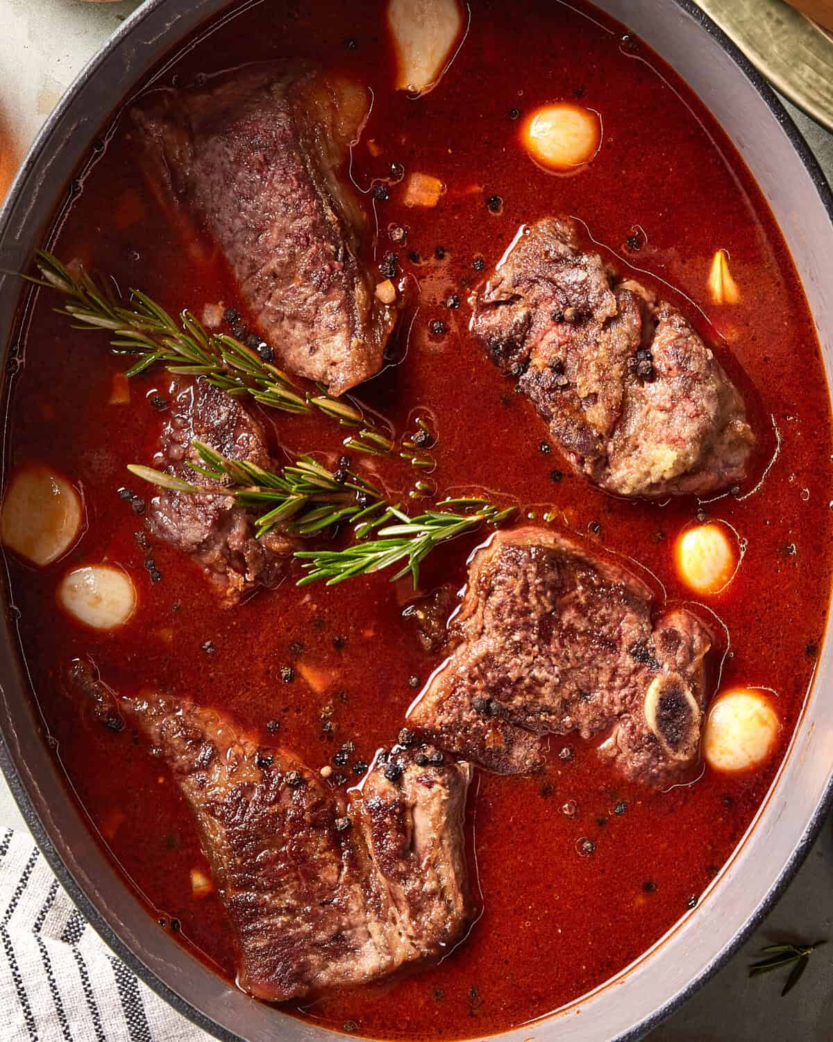 Chunks of braised beef in a rich red sauce, garnished with sprigs of rosemary and whole garlic cloves, simmering in a pot.