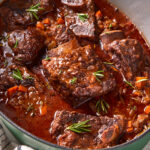 Braised short ribs in a rich, red sauce with chopped carrots, onions, and rosemary sprigs, served in a green Dutch oven on a striped towel.
