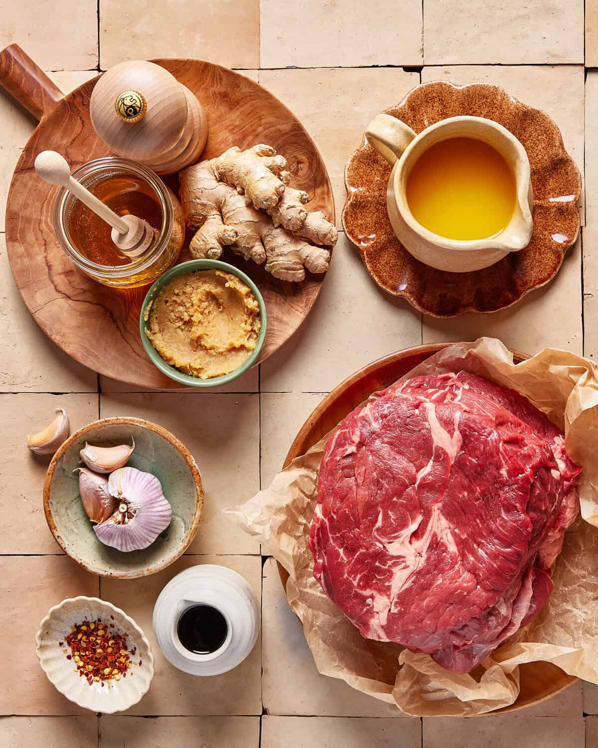 A flat lay of cooking ingredients on a tiled surface, including a raw marbled steak, fresh ginger, garlic cloves, honey, ground ginger, pepper grinder, chili flakes, soy sauce, and a small pitcher of oil.