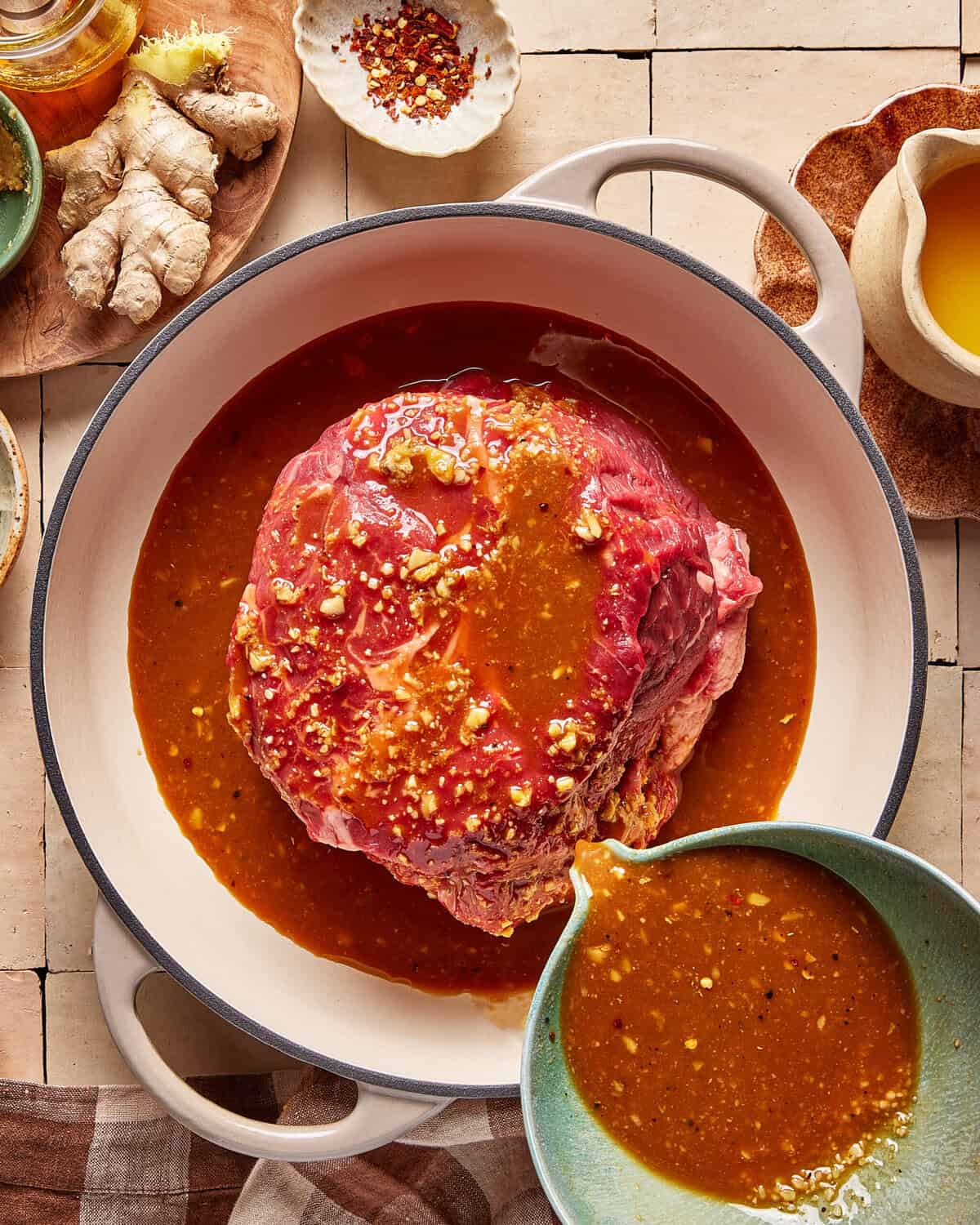 A large raw beef roast sits in a pot, covered with a brown marinade. A bowl pours more marinade over it. Ginger, crushed red pepper, and a cloth napkin are arranged nearby on a tiled surface.
