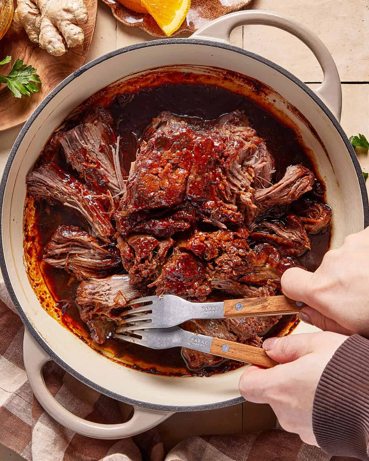 Hands using two forks to shred a large, cooked piece of beef in a round, enameled cast iron pot filled with rich, dark sauce. Fresh ginger and herbs are visible on the side.