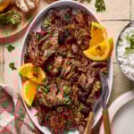 A platter of cooked shredded beef garnished with parsley and orange slices, served alongside a bowl of white rice. A checkered napkin, forks, ginger root, and fresh parsley are nearby on a tiled surface.