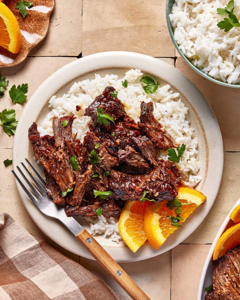 A plate of shredded beef served over white rice, garnished with fresh parsley and orange slices. A fork rests on the plate, with a bowl of white rice and a napkin nearby.
