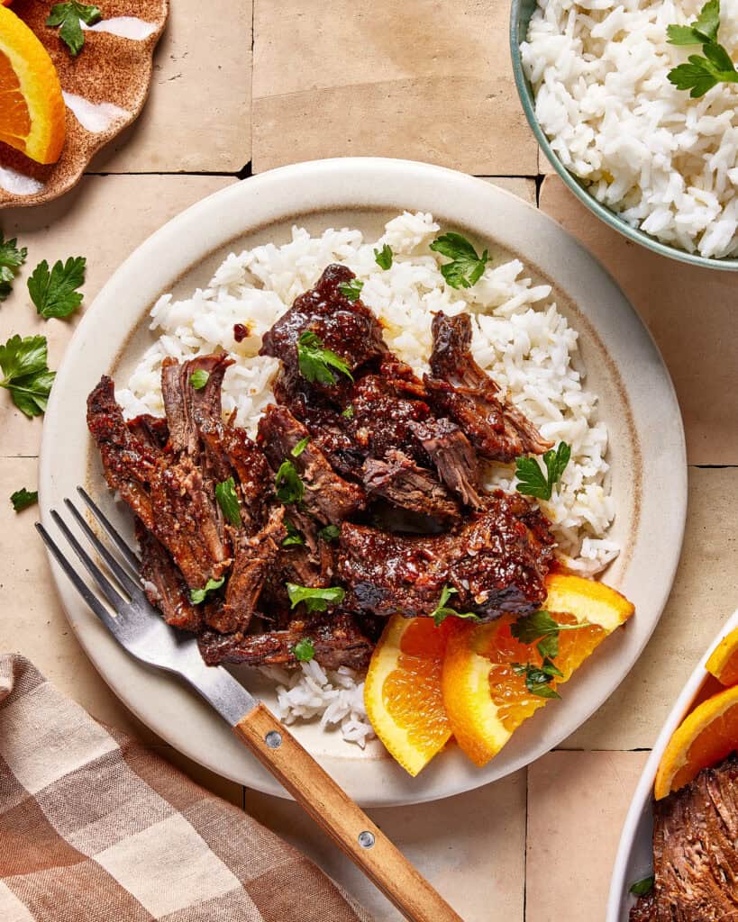 A plate of shredded beef served over white rice, garnished with fresh parsley and orange slices. A fork rests on the plate, with a bowl of white rice and a napkin nearby.