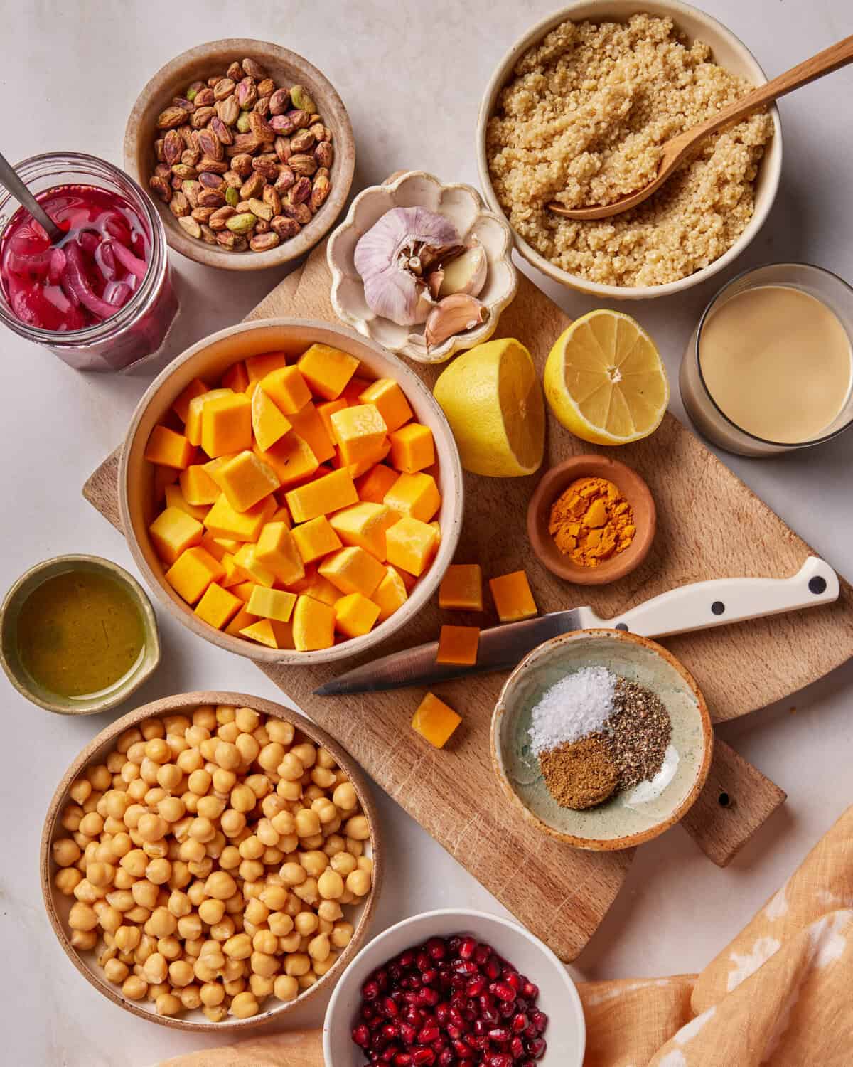 A variety of ingredients in bowls on a table, including cubed butternut squash, chickpeas, quinoa, pistachios, pomegranate seeds, lemon, pickled onions, garlic, spices, olive oil, and tahini, with a knife on a cutting board.